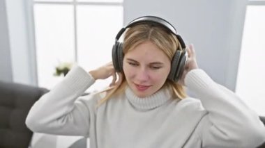 Sarışın ve kulaklıklı, rahat, beyaz bir kadın rahat bir oturma odasında kanepeli müzikten hoşlanıyor..