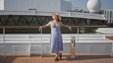 Yazlık elbisesi ve şapkası olan genç bir kadın okyanus zeminine karşı bir yolcu gemisinde gülümsüyor..