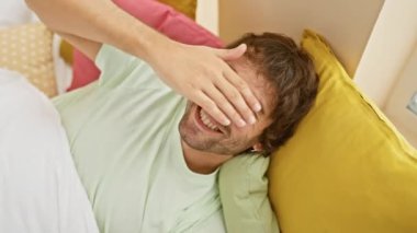 Küstah, genç, beyaz bir adam, şakacı bir şekilde gözlerini kapatıyor, rahat bir yatak odasında sürpriz bir gülümseme sergiliyor, pijama giyiyor, kör bir eğlenceyi ima ediyor.