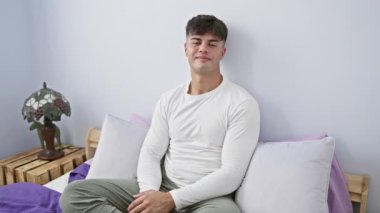 Neşeli genç İspanyol adam yatak odasında poz veriyor, pijamalarıyla güvenle gülümsüyor, kollarını kavuşturup yatağa uzanıyor.