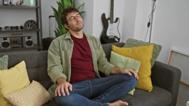Rahat bir adam gri bir kanepede kapalı gözleri ile oturan rahat oturma odasında, renkli yastıklar ve arka planda bir gitar ile çevrili.