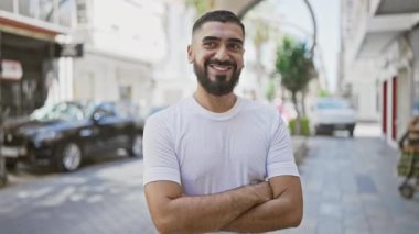 Güler yüzlü, sakallı bir adam güneşli bir caddede kolları bağlı duruyor..