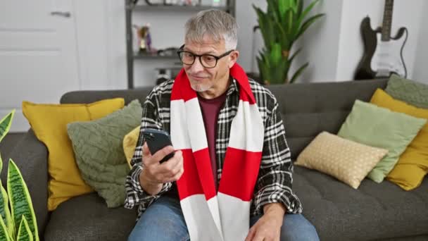 スマートフォンを使用してスカーフを身に着けている中年の灰色の髪の男性は指でOkサインをし 自宅で優れたシンボルを微笑みます — ストック動画