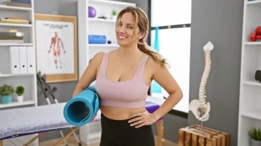 Rehabilitasyon kliniğinde yoga minderi tutan kendine güvenen bir kadın. Arka planda iskelet modeli ve fizyoterapi ekipmanları var..