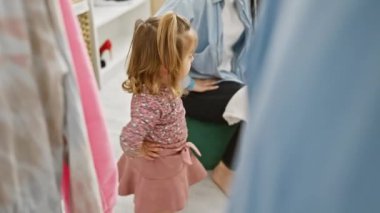 Pembe etekli genç bir kız modern bir soyunma odasında kıyafetlerin arasında annesine sarılıyor..