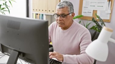 Gözlük takan olgun işadamı kapalı ofis ortamında bilgisayara odaklanıyor..