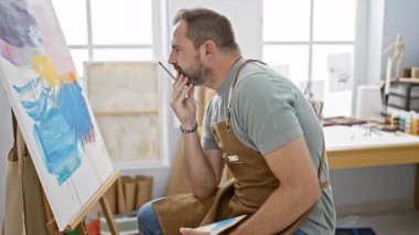 Önlüklü düşünceli bir adam parlak bir sanat stüdyosunda tuvali inceliyor..