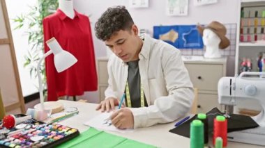 Genç bir adam dikiş malzemeleri ve mankenlerle çevrili bir terzi atölyesinde moda tasarımları çiziyor..