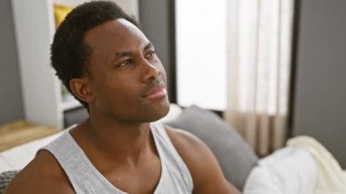 Genç Afro-Amerikalı adam pijama giymiş gülümseyip yan tarafa bakıyor ve düşünüyor. yatak odasında