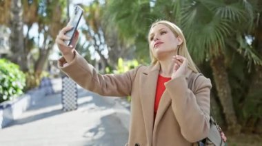Palto giymiş genç bir kadın güneşli bir patikada selfie çekiyor.