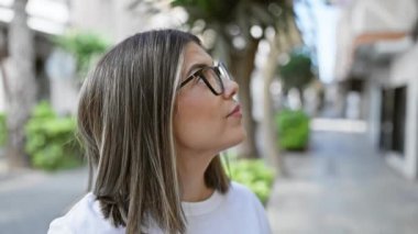 Gözlüklü, kendine güvenen bir İspanyol kadın şehir ortamında gülümsüyor, pozitifliği ve gündelik tarzını yansıtıyor..