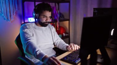 Kulaklık takan İspanyol sakallı adam geceleri neon ışıklı bir oyun odasında bilgisayara konsantre oluyor..