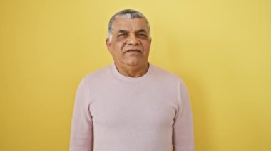 Şok olmuş orta yaşlı, gri saçlı ve süveter giyen Latin bir adam, sarı bir arka planda şaşırtıcı bir korku ifadesi ve şaşkın bir sevinçle tek başına duruyor..