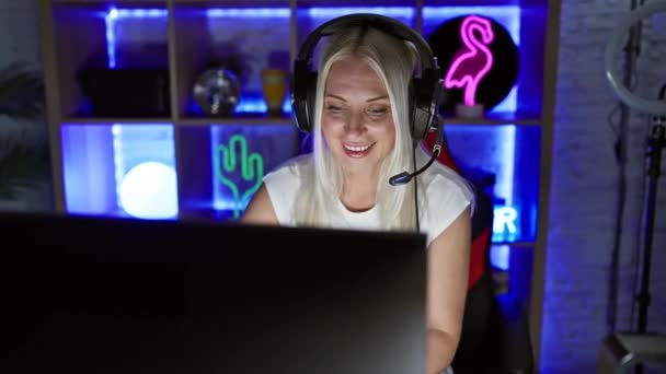 兴奋的年轻金发女人带着耳机笑着 晶莹无瑕的牙齿笑着 完全沉浸在电脑游戏中 在家里的游戏室里流露出喜悦 — 图库视频影像
