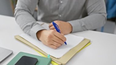 Ofiste yazan Asyalı genç bir adam. Etrafı teknoloji ve belgelerle çevrili.