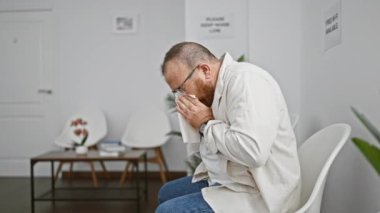 Endişeli, orta yaşlı, gözlüklü, hastalıktan muzdarip beyaz bir adam, elinde kağıt peçeteyle oturma odasında oturuyor, stresli bir şekilde hapşırıyor.