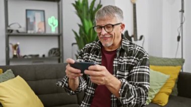 Gözlüklü ve ekose gömlekli neşeli bir son sınıf öğrencisi modern bir oturma odasında akıllı telefon kullanıyor..