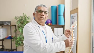 Beyaz önlüklü olgun bir adam rehabilitasyon kliniğinin terapi odasında omurga modelini inceliyor..
