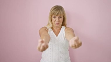 Cheerful middle age blonde woman excited yet with a bad attitude, showing a 'fuck you' sign with her middle finger over an isolated pink background