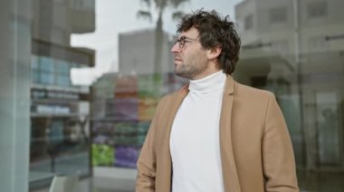 Gözlüklü ve sakallı sıradan bir genç arka planda ofis binaları olan bir caddede dikiliyor.