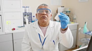 Laboratuvar önlüğü ve güvenlik gözlüğü takan olgun bir adam iyi donanımlı bir laboratuarda kimyasal bir örneği inceliyor.