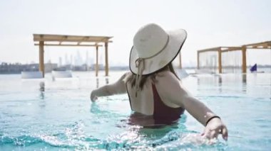 Mayo giymiş neşeli bir kadın, arka planda açık bir gökyüzünün altında şehir silueti olan lüks bir havuzda dinleniyor..