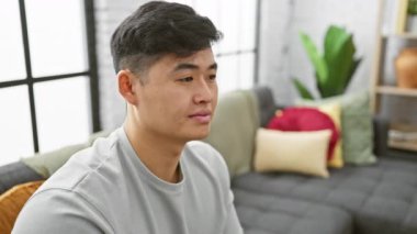 Düşünceli genç Asyalı bir adam modern bir oturma odasında tek başına oturuyor..