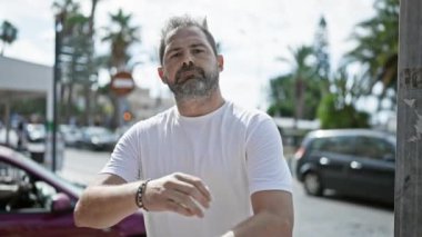 Gri saçlı, çapraz kollu, kenar mahallede duran kendine güvenen olgun bir adam..