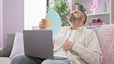 Gri saçlı, olgun sakallı bir adam evinde dizüstü bilgisayarı üzerinde çalışırken el yelpaze kullanıyor ve rahat bir atmosfer yayıyor..