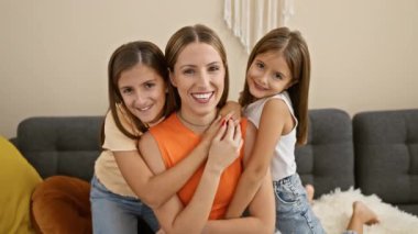 Gülümseyen bir kadın sıcak bir oturma odasında iki mutlu kızını kucaklıyor, sıcaklığı ve aileyi resmediyor..