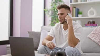 Sıradan kıyafetlerle sakallı odaklanmış genç bir adam evde dizüstü bilgisayarla çalışırken telefonla konuşuyor.