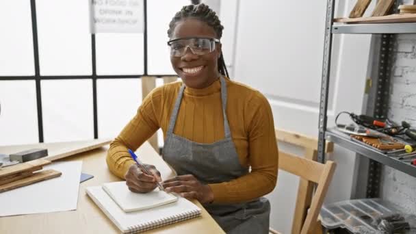 Szczęśliwa Kobieta Warkoczach Okularach Bezpieczeństwa Pisze Notatniku Ławce Stolarskiej — Wideo stockowe