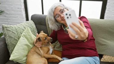 Gülümseyen yaşlı bir kadın, köpeğiyle oturma odasındaki rahat bir kanepede selfie çeker..