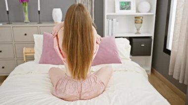 Genç bir kadın yatak odasında bir yatakta oturur, sırtını kameraya dayar, rahat, modern bir iç mekanda..