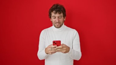 Ağzını eliyle kapatan, konuşamayan, kırmızı izole edilmiş arka planda akıllı telefon kullanan, şoke olmuş genç bir İspanyol. gizli sürpriz hareketi, korku içinde dilsiz, yakışıklı sakallı yüz ifadesini gizlemek.