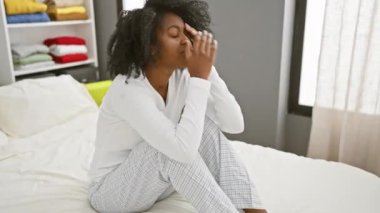 Stresli bir Afrikalı Amerikalı kadın yatak odasında bir yatakta oturuyor, yorgun ve endişeli görünüyor.