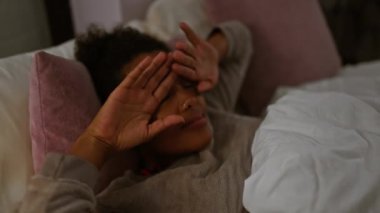 Yatak odasında gevşeyen kıvırcık saçlı genç bir kadın, gözlerini ışıktan koruyor, sıcak ve samimi bir ortamda yakalanmış..