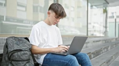 Kampüs merdivenlerinde oturan genç bir adam yanında sırt çantasıyla dizüstü bilgisayar kullanıyor..