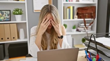 Modern bir ofis ortamında kendini yorgun hisseden stresli sarışın bir kadın başını ellerinin arasına almış dinleniyor..