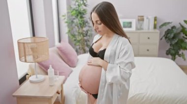 Düşünceli genç bir İspanyol kadın, rahat, aydınlık bir yatak odasında yatağın yanında dururken hamile karnını nazikçe kucaklıyor..