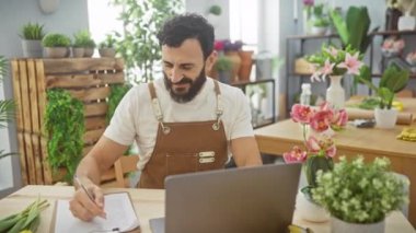 Çiçekçide gülümseyen sakallı bir adam laptopun yanına notlar yazar. Etrafı yemyeşil bitkiler ve renkli çiçeklerle çevrilidir..