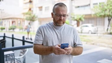 Neşeli, orta yaşlı, gözlüklü, sakallı, neşeli bir şekilde cep telefonunu kullanan, çevrim içi bir sohbete dalmış, hareketli bir şehir caddesinde gülümseyen beyaz bir adam.