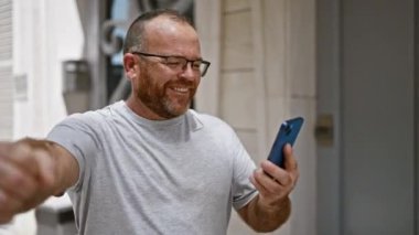 Neşeli, gözlüklü, sakallı, orta yaşlı, beyaz bir adam canlı, şehir sokağında telefonundan bangır bangır müzik dinliyor