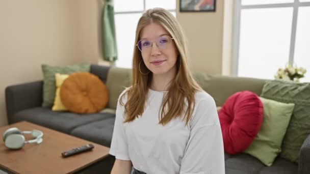 一个戴着眼镜的快乐的金发女人在一个舒适的客厅里摆姿势 房间里有沙发和现代装潢 — 图库视频影像