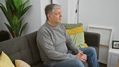 Modern bir oturma odasında gri bir kanepede otururken sırtı ağrıyan olgun gri saçlı bir adam.