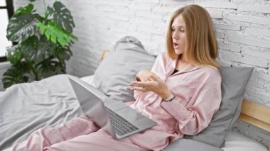 Pembe pijamalı sarışın bir kadın modern bir yatak odasında yatarken laptopunda bir video görüşmesi yapıyor.