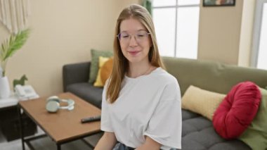 Gülümseyen, gözlüklü, sıradan giyinmiş, modern dekorlu, rahat bir oturma odasında rahatlayan sarışın bir kadın..