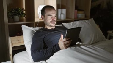 Geceleri rahat bir odada tablet kullanan gülümseyen sakallı adam.