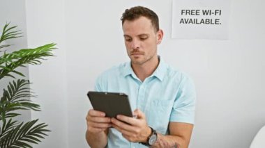 Beyaz bekleme odasında tablet kullanan açık mavi tişörtlü yakışıklı İspanyol adam. Yanında da ücretsiz wi-fi işareti var..