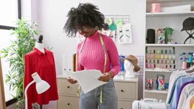 Gözlüklü ve mezuralı Afrikalı kadın mankenli ve dikiş makineli iyi donanımlı bir atölyede bir desen okuyor..